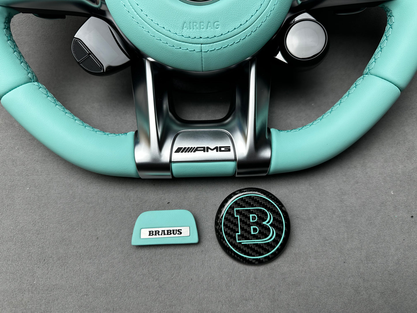 Close-up of Brabus Tiffany Blue flat emblem and round carbon B emblem on a Mercedes steering wheel