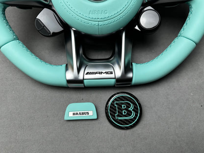 Close-up of Brabus Tiffany Blue flat emblem and round carbon B emblem on a Mercedes steering wheel